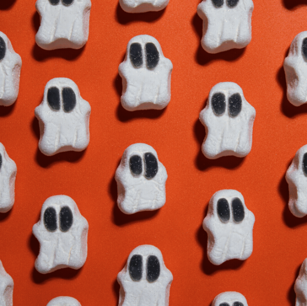 White ghost-shaped marshmallows with black eyes arranged in a repeating pattern on a bright orange background.